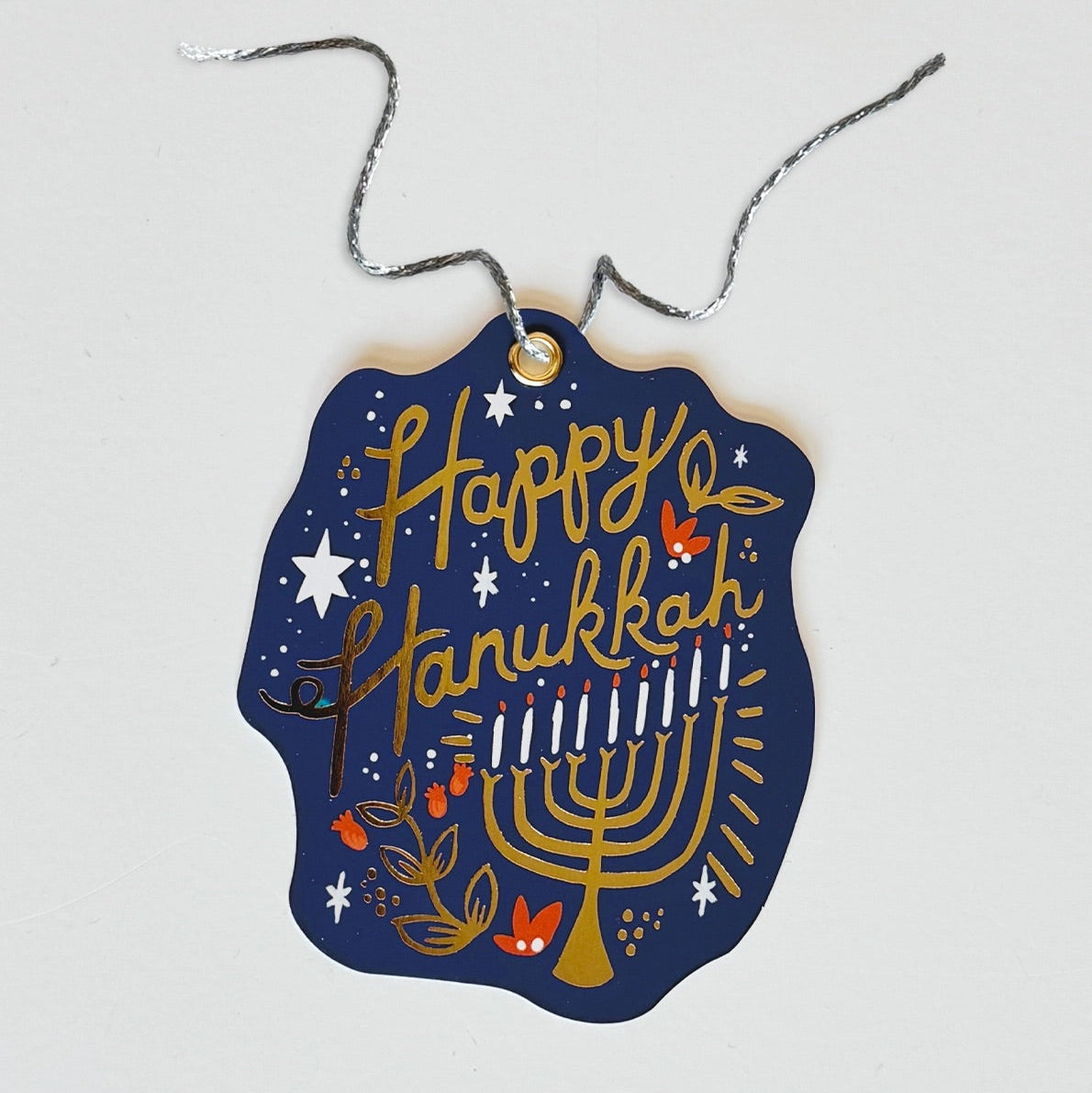 A navy blue gift tag with a gold menorah, stars, and the words 'HAPPY HANUKKAH' written in a bold, decorative font. It includes a loop for attaching a twine.