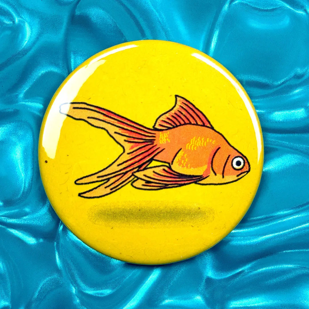 Button badge with a goldfish design on a yellow background, placed on a blue textured surface.