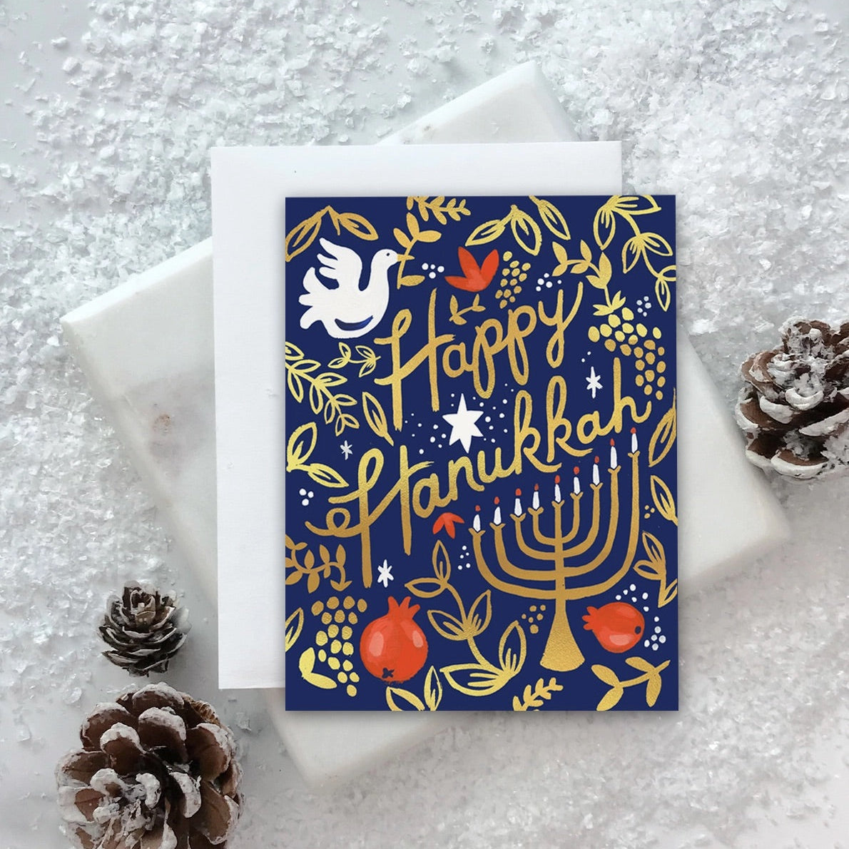 A folded Hanukkah-themed greeting card with gold foil details, featuring a pomegranate and floral design, placed on a white surface with pine cones nearby.