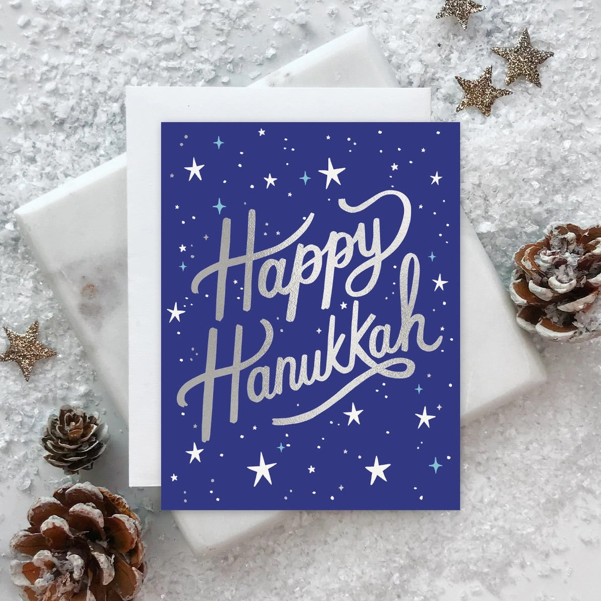 A blue greeting card with silver stars and the text 'HAPPY HANUKKAH' in a bold script, placed on a white surface with decorative silver foil and pine cones around it.