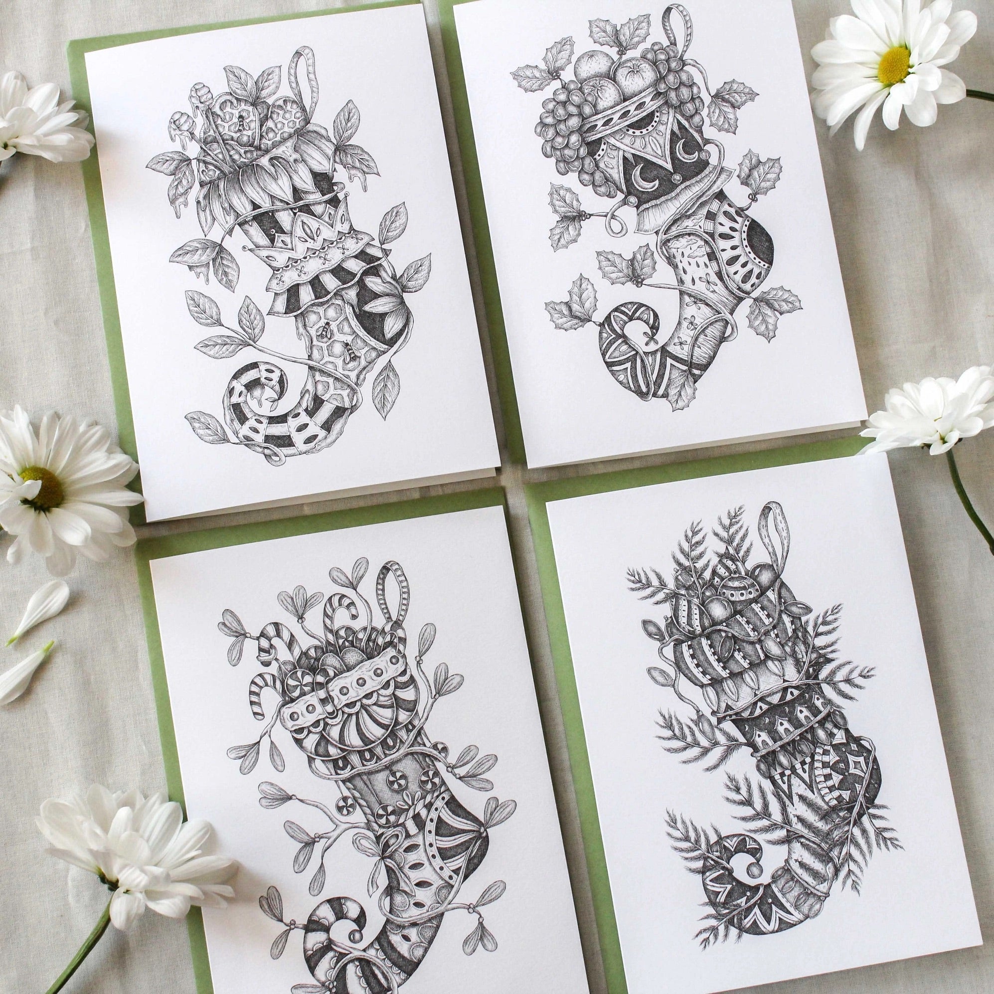A set of four black and white holiday cards with detailed and artistic illustrations of stockings, displayed on a surface with some white flowers around them.