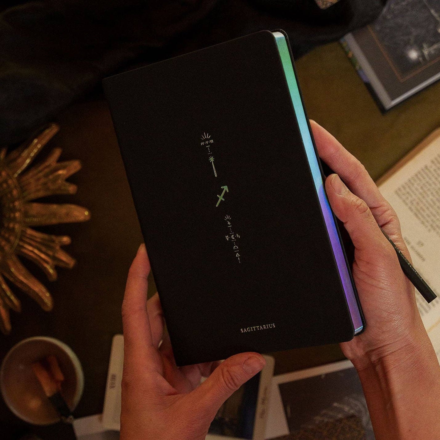 Magic of I - Zodiac Journal - Sagittarius - Person holding a black notebook with colorful edges on a table.