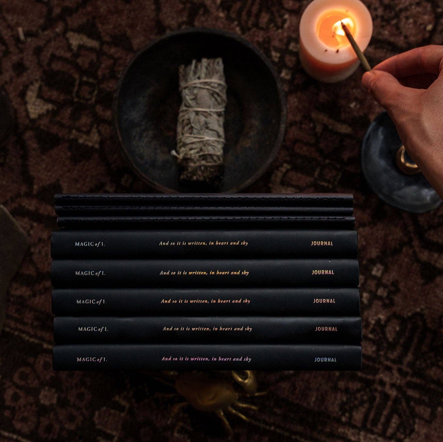 Candle being lit next to a bundle of sage on a textured surface with black books displaying text.